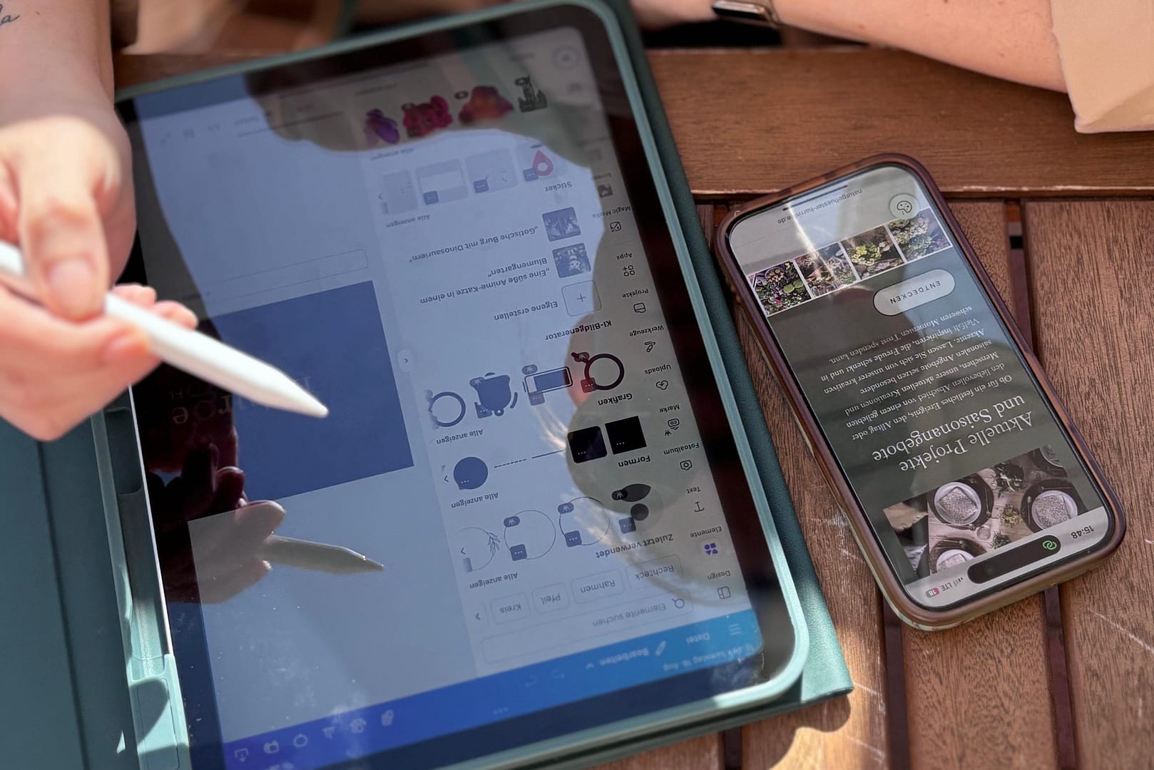 A close-up of a person's hand holding a stylus, pointing at a tablet screen displaying a design interface, with a smartphone nearby showing a floral arrangement website.